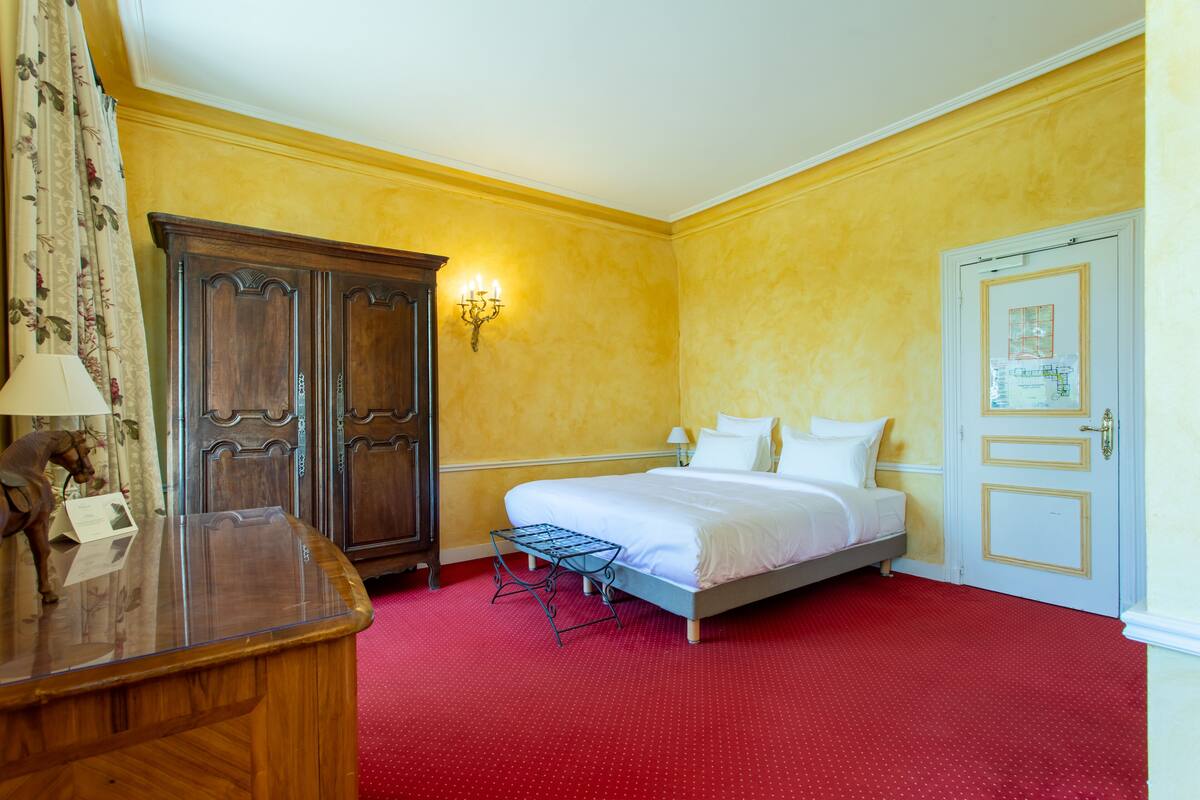 A hotel room with a bed, a wooden desk, a lamp, a wall-mounted light fixture, and a door with a small window.