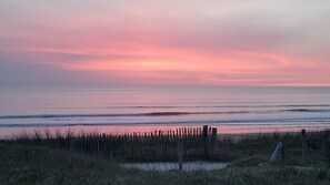 A beach with a wooden fence in the foreground and a calm sea under a pink and purple sky.