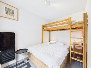 Uma cama de solteiro com estrutura de madeira, uma mesa pequena e uma cadeira num quarto com paredes brancas.