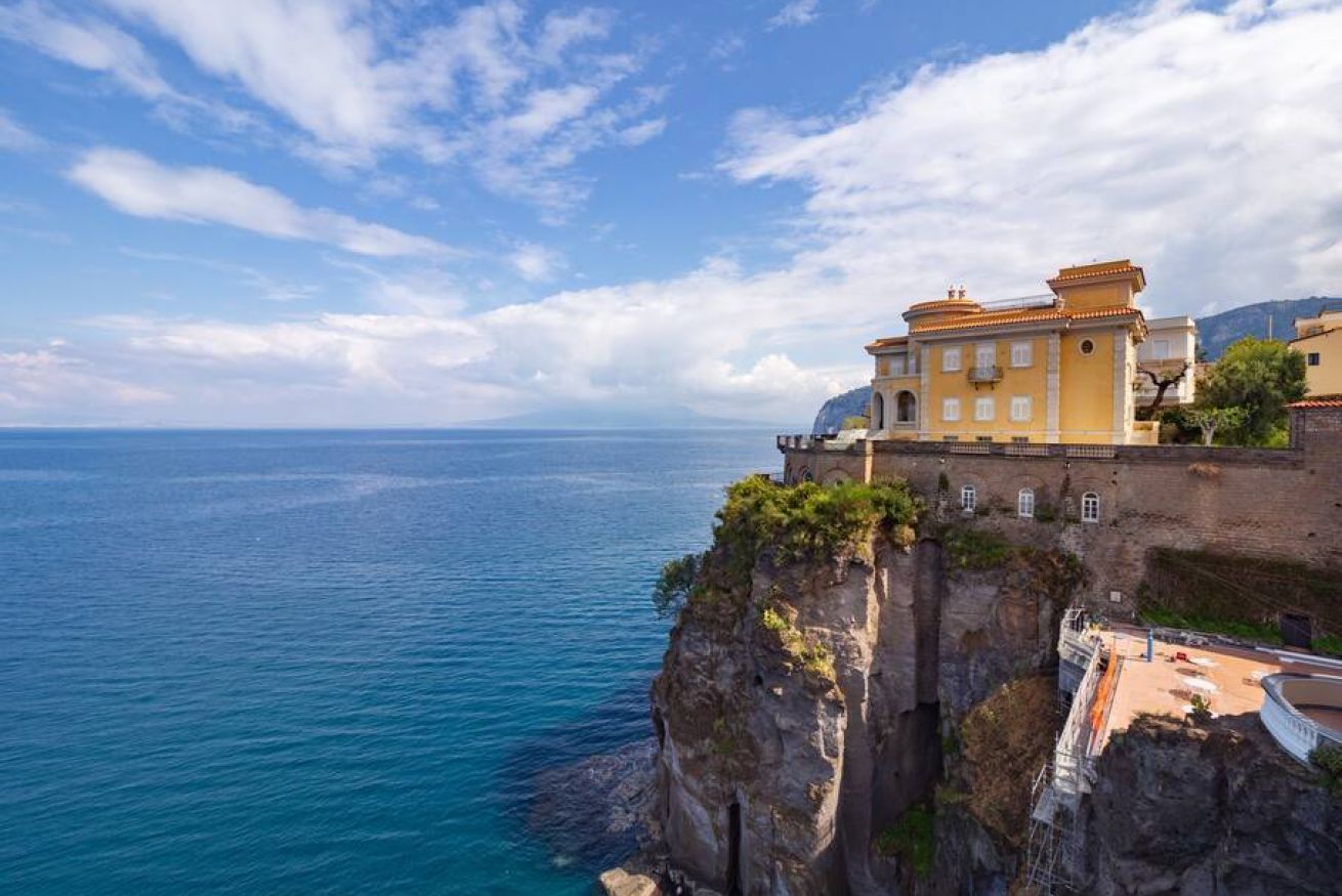Un bâtiment situé sur la côte, doté d’un balcon donnant sur la mer, sous un ciel dégagé, et adossé à une falaise rocheuse.