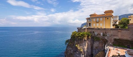Un bâtiment situé sur la côte, doté d’un balcon donnant sur la mer, sous un ciel dégagé, et adossé à une falaise rocheuse.