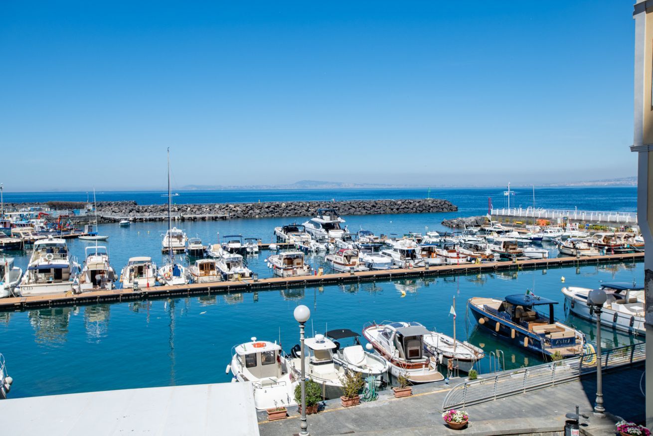 Un port de plaisance où sont amarrés différents types de bateaux, un ciel dégagé et une digue rocheuse en arrière-plan.
