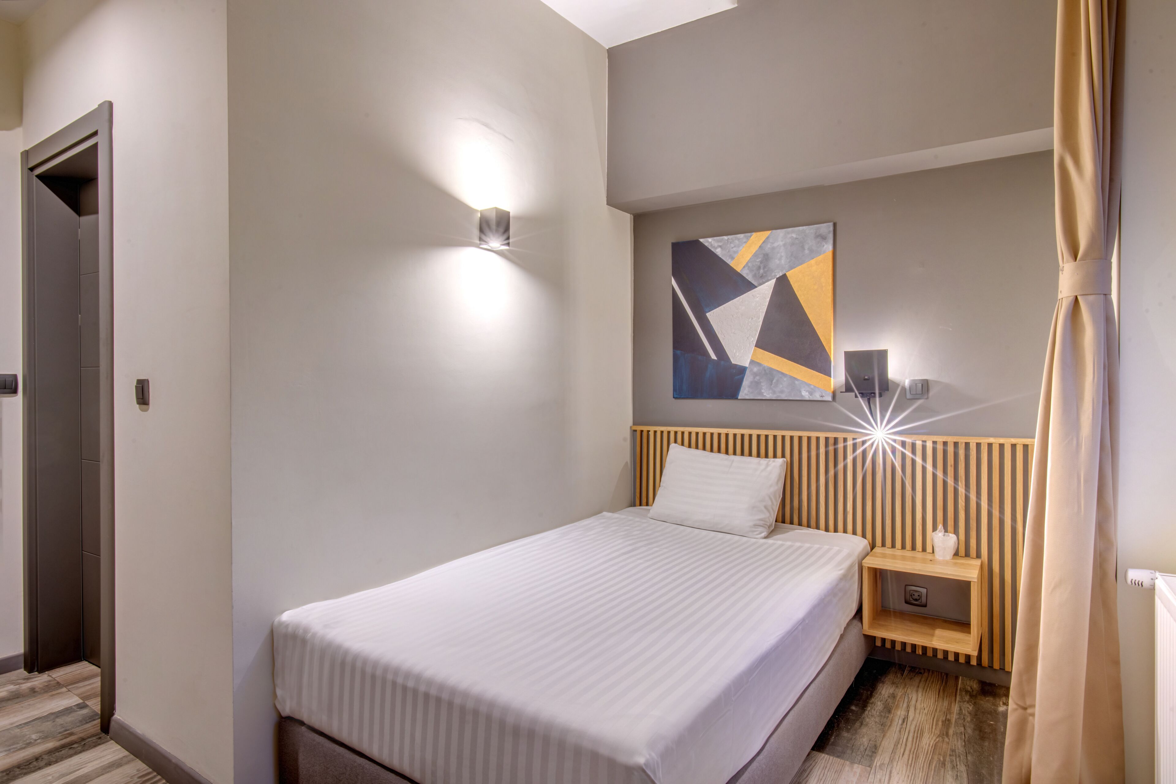 A modern hotel room with a neatly made bed, a bedside table with a lamp, and a geometric artwork on the wall.