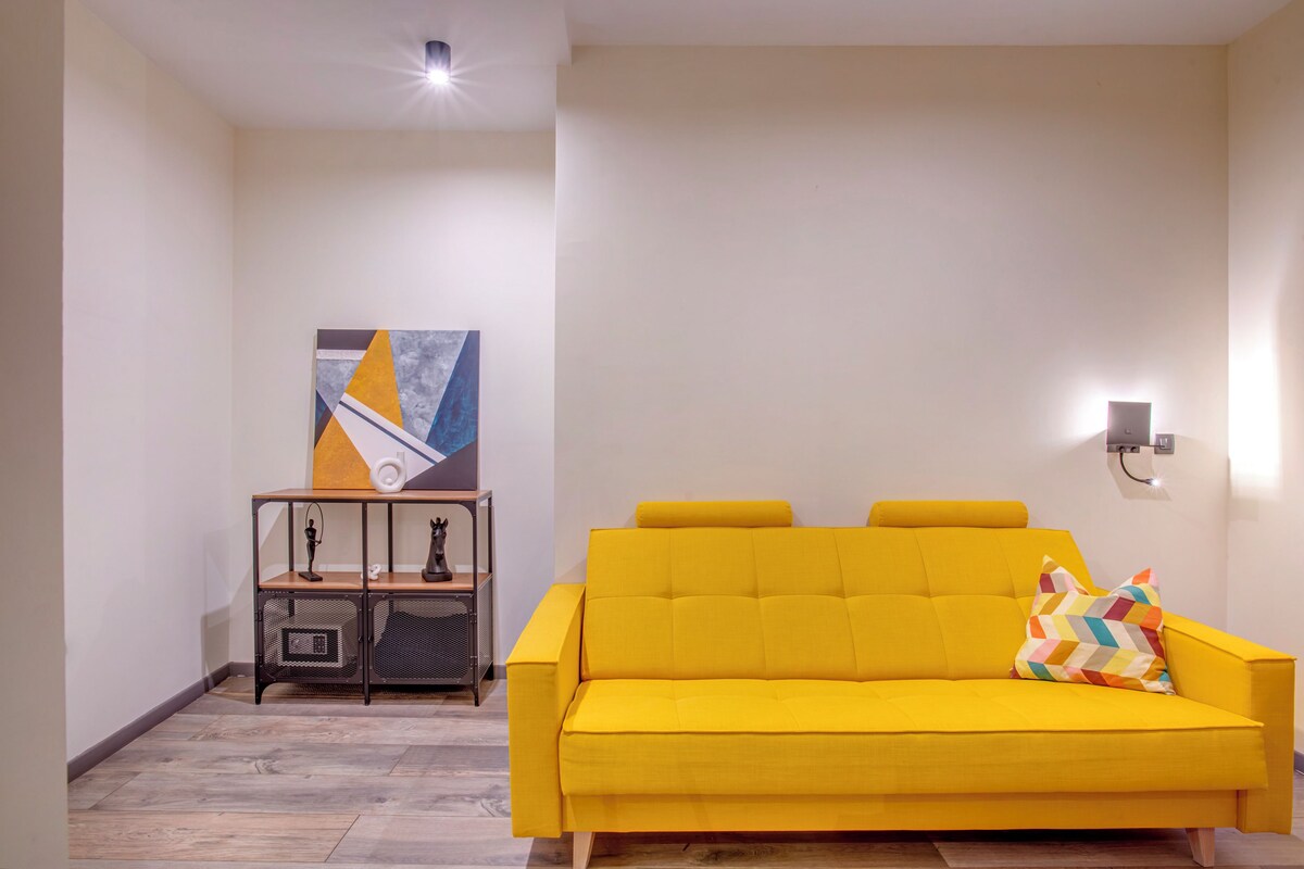 Un sofá amarillo brillante con un cojín colorido, una mesita auxiliar con una lámpara y un aplique de pared.