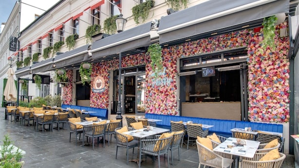 Ein Straßencafé mit Korbtischen und -stühlen, einer blauen Bank und einer Wanddekoration mit Blumen.