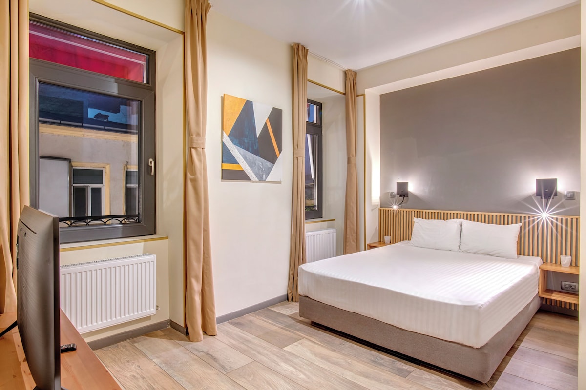 Un dormitorio con una cama grande, una mesita de noche con lámpara, una ventana con cortinas, un radiador y una obra de arte colgada en la pared.