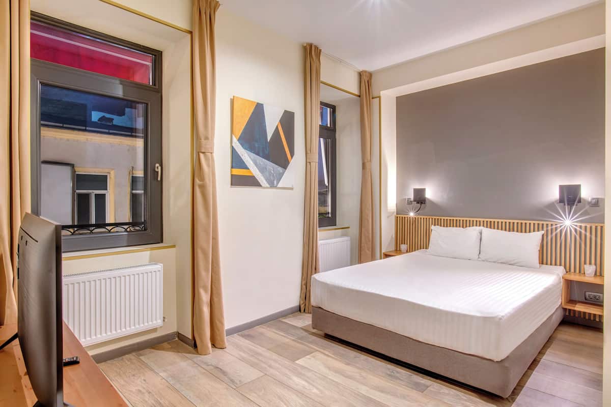 A bedroom with a large bed, a bedside table with a lamp, a window with curtains, a radiator, and a wall-mounted artwork.