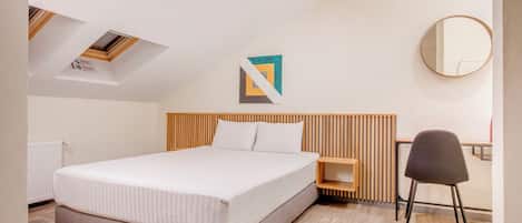 A modern bedroom with a large bed, a wooden headboard, a bedside table, a chair, a round mirror, and a geometric artwork on the wall.