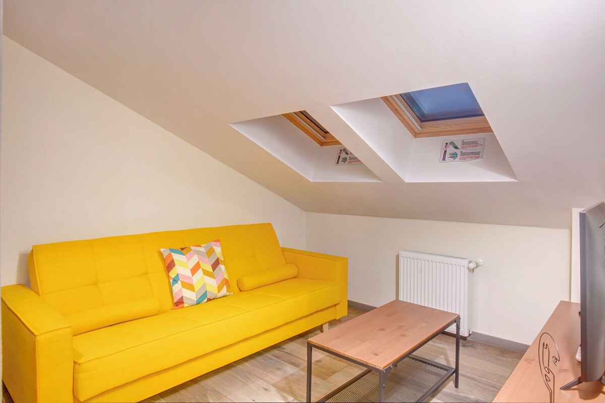 Un sofá amarillo brillante con un cojín colorido, una mesa de centro de madera y un radiador en la pared.