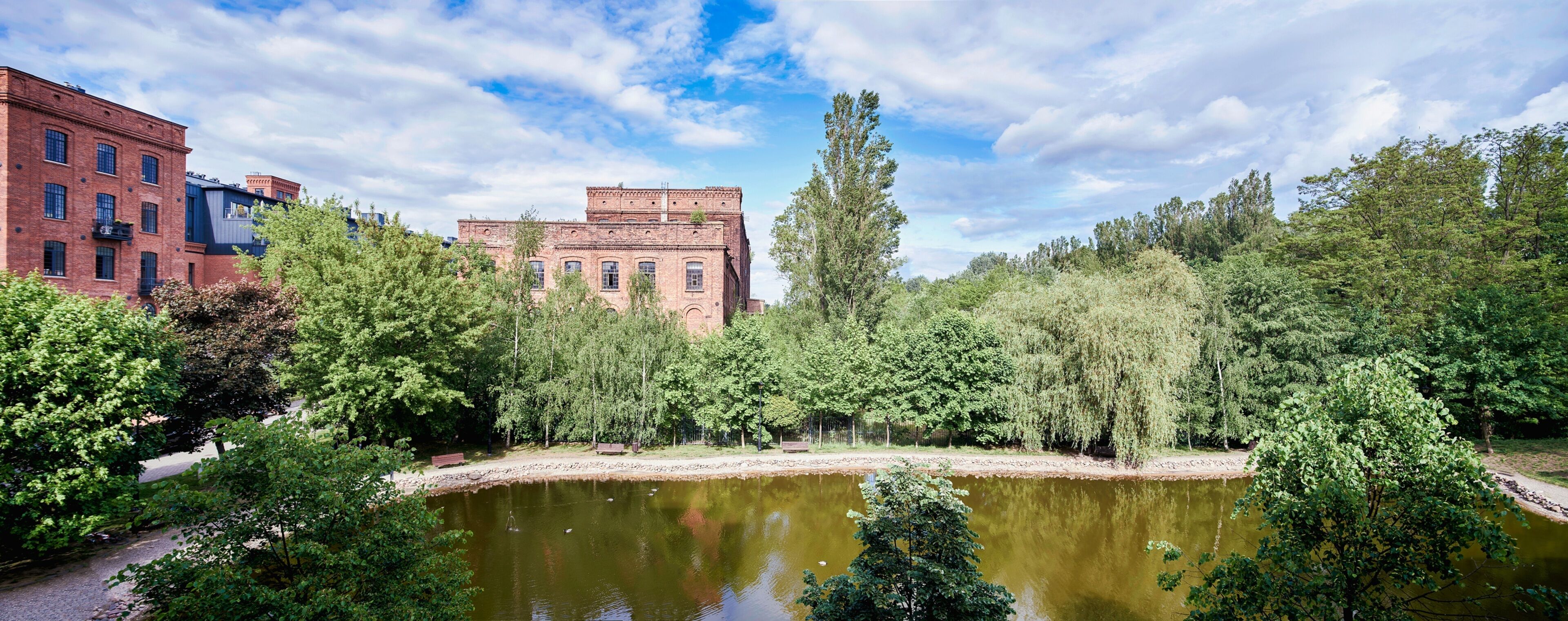 Un bâtiment historique à la façade en briques et aux grandes fenêtres, entouré d’une végétation luxuriante et d’un petit étang.