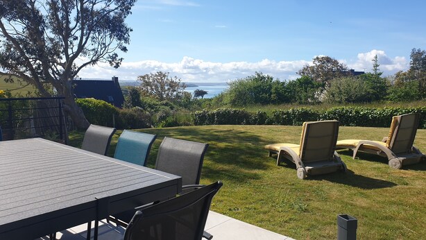 Une terrasse avec une table et des chaises, une vue sur la mer et un espace verdoyant avec des arbres.