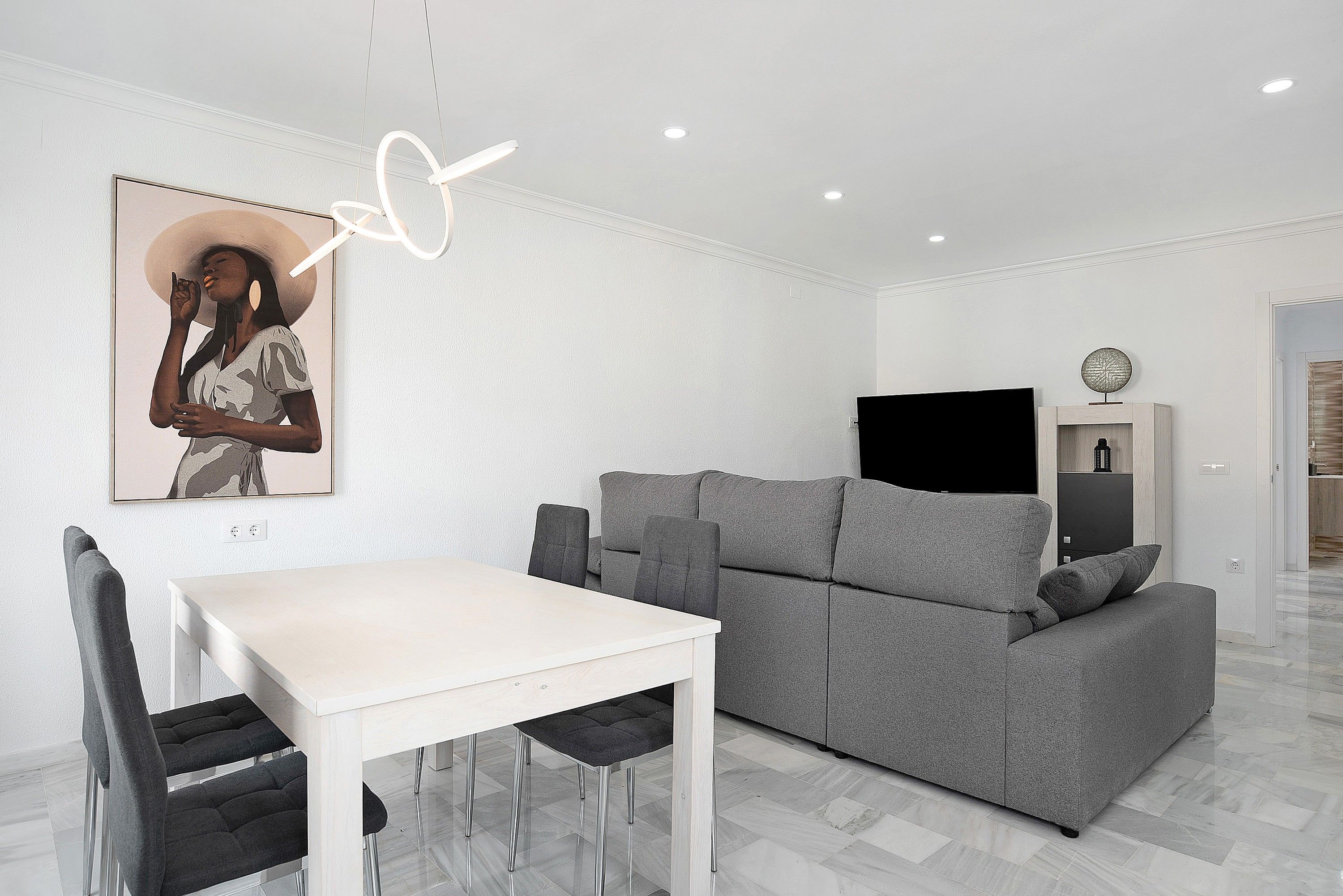 Una sala moderna con un sofá gris, una mesa de comedor blanca y un televisor de pantalla plana negra.