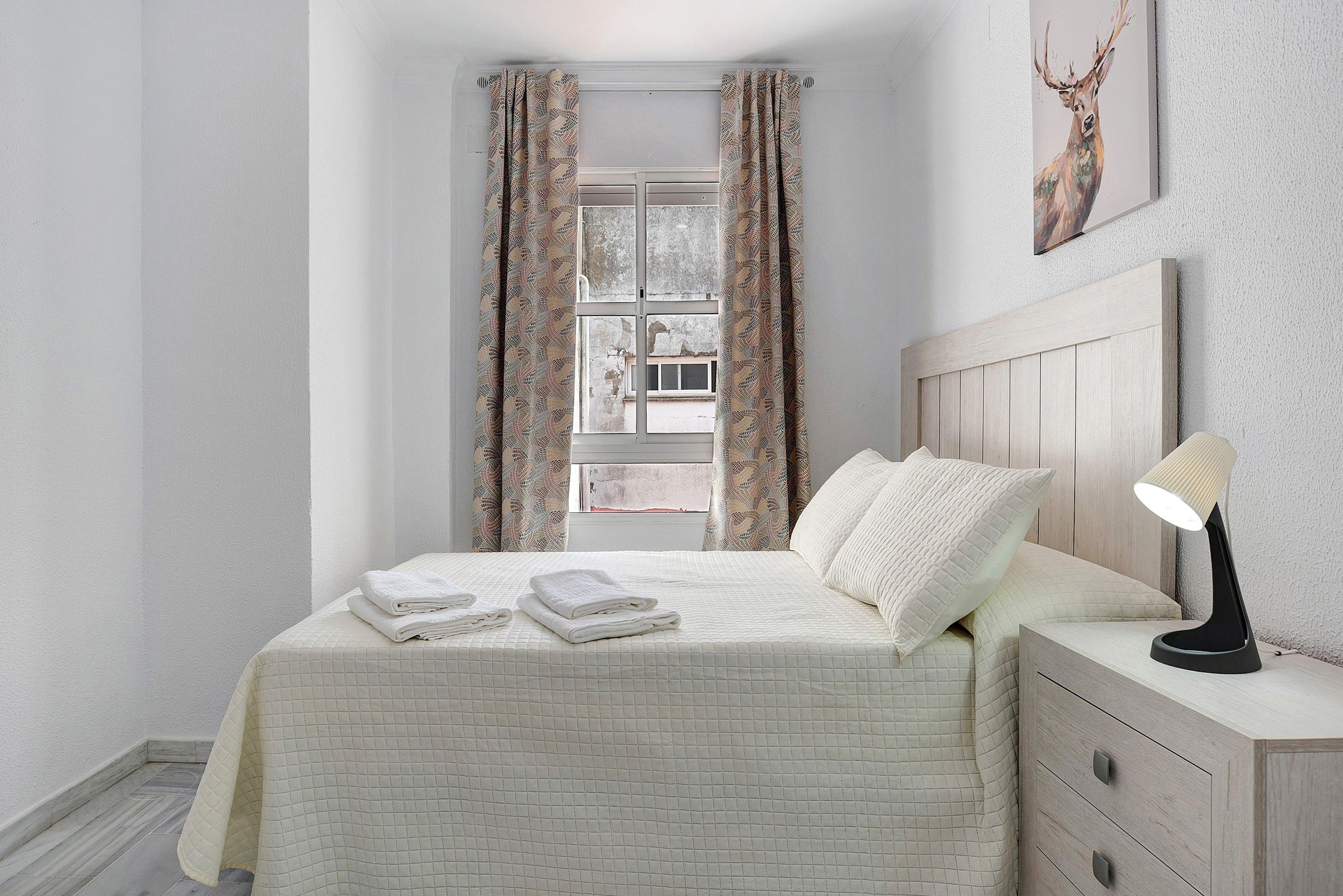 Un dormitorio con cama, mesita de noche, lámpara, ventana con cortinas y un cuadro en la pared.