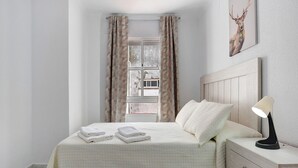 Une chambre à coucher avec un lit, une table de chevet, une lampe, une fenêtre avec des rideaux et un tableau au mur.