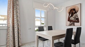 Un coin repas avec une table de couleur claire et des chaises foncées, une fenêtre avec des rideaux blancs et un luminaire fixé au mur.