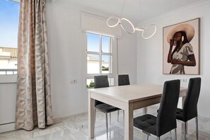Een eetruimte met een lichtgekleurde tafel en donkere stoelen, een raam met witte gordijnen en een wandlamp.