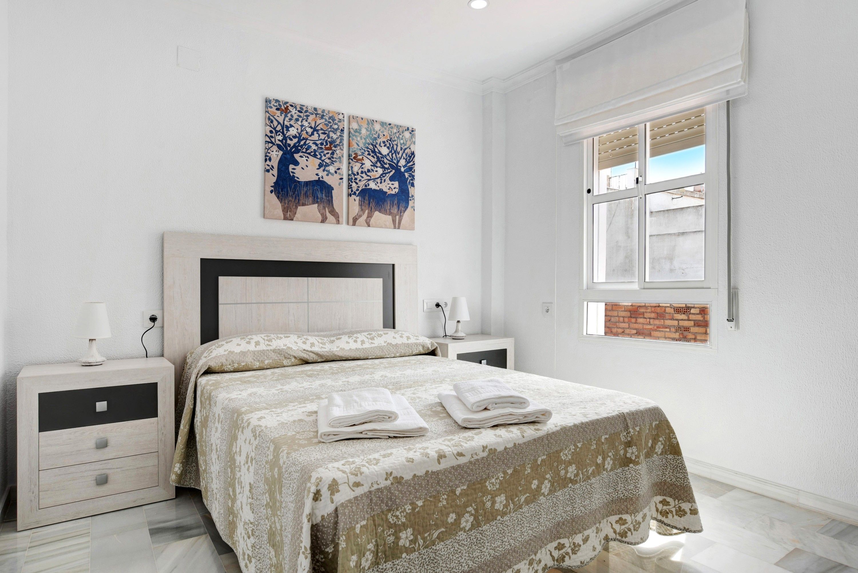 Un dormitorio con cama, mesitas de noche, una ventana y cuadros en la pared.