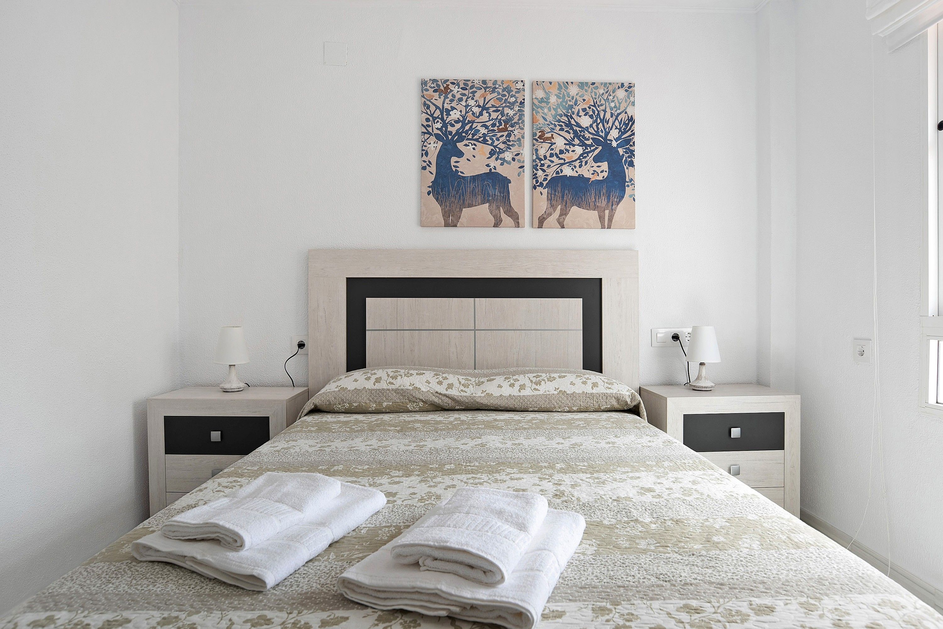 Una cama bien hecha con dos toallas dobladas, un cabecero, dos mesitas de noche con lámparas y dos cuadros colgados en la pared.