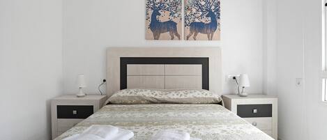 A neatly made bed with two folded towels, a headboard, two bedside tables with lamps, and two wall-mounted paintings.