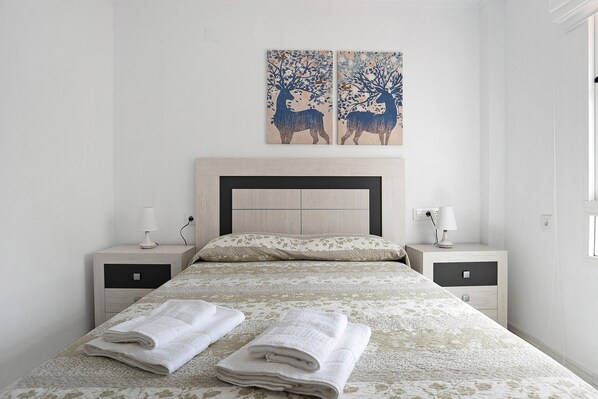 A neatly made bed with two folded towels, a headboard, two bedside tables with lamps, and two wall-mounted paintings.