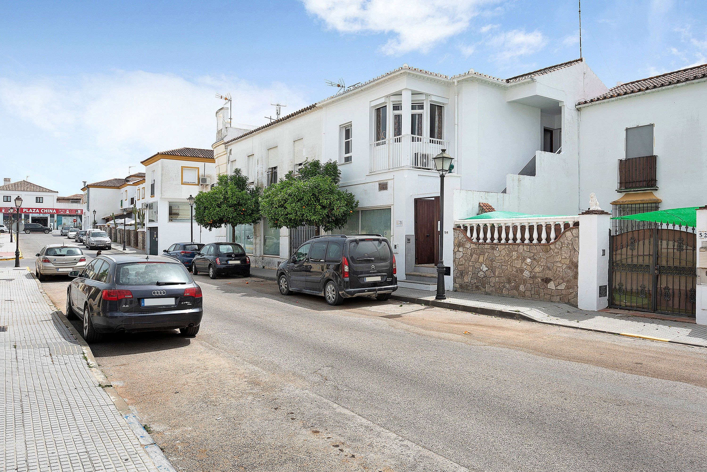 Vista de la calle con coches aparcados, un edificio blanco con balcón y un muro de piedra con una reja.