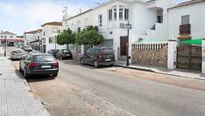 Une vue de rue avec des voitures garées, un immeuble blanc doté d’un balcon et un mur de pierre avec un portail.