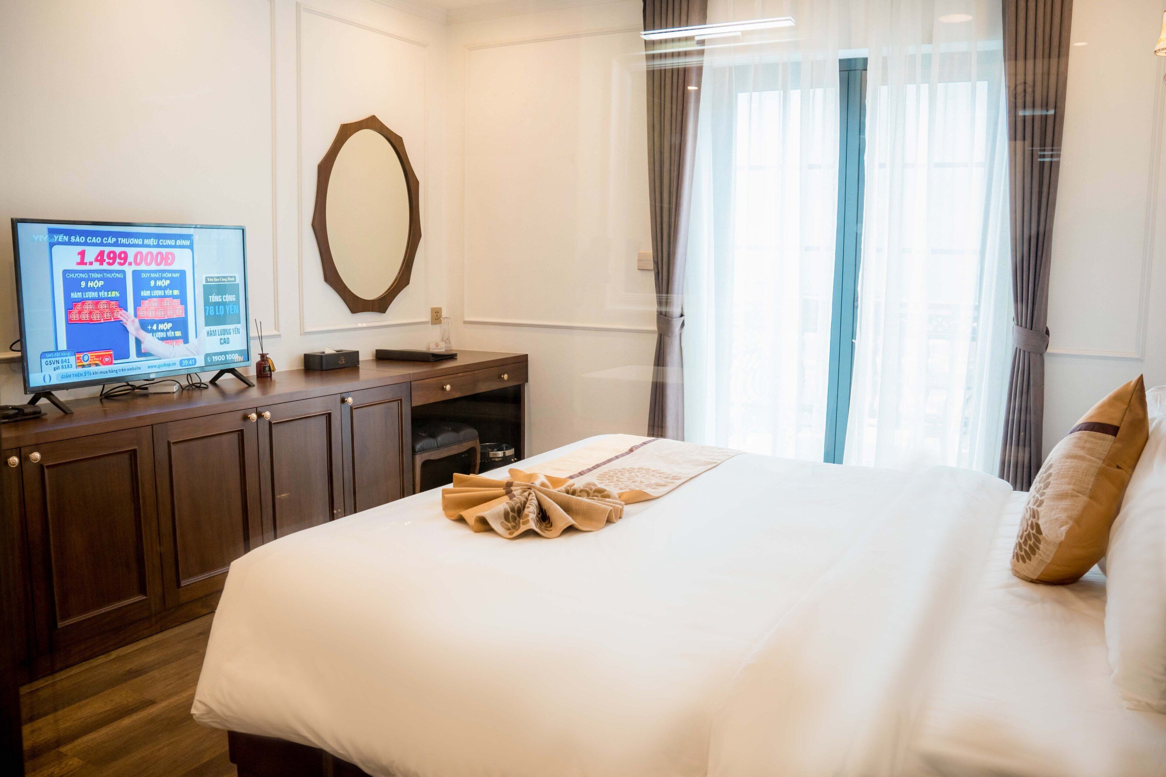 A hotel room with a bed, a TV on a wooden desk, a mirror, and a window with curtains.