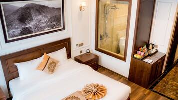 Habitación de hotel con una cama grande, un cuadro colgado de un paisaje montañoso, ducha con mampara de vidrio y un tocador pequeño con artículos de aseo.
