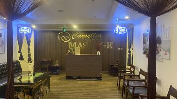 A reception area with a wooden reception desk, a wall-mounted sign reading "Camellia Hotel Massage," and a tiled floor.