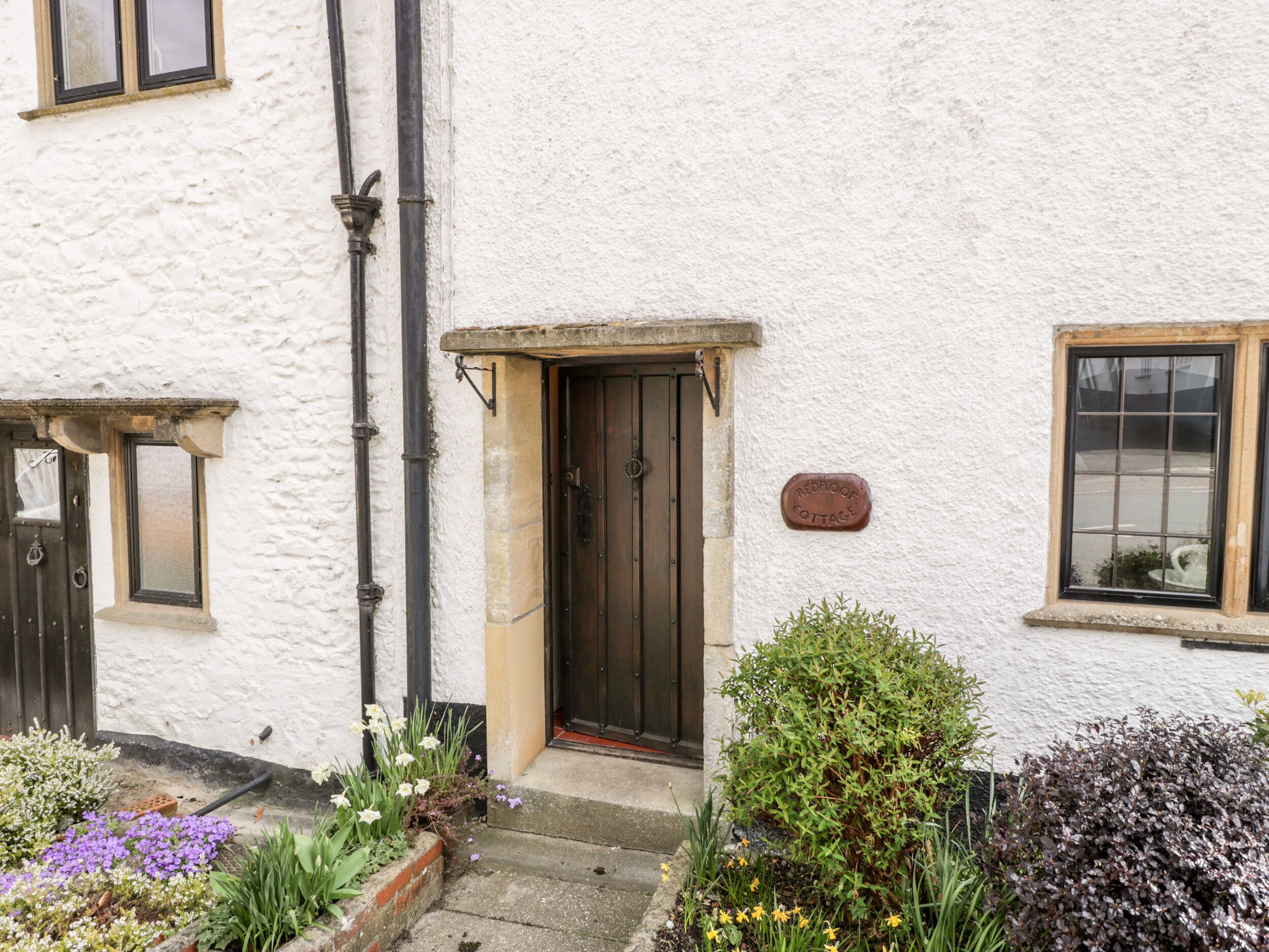 A white building with a dark wooden door, a small garden with flowers, and a sign reading "Redrock Cottage".