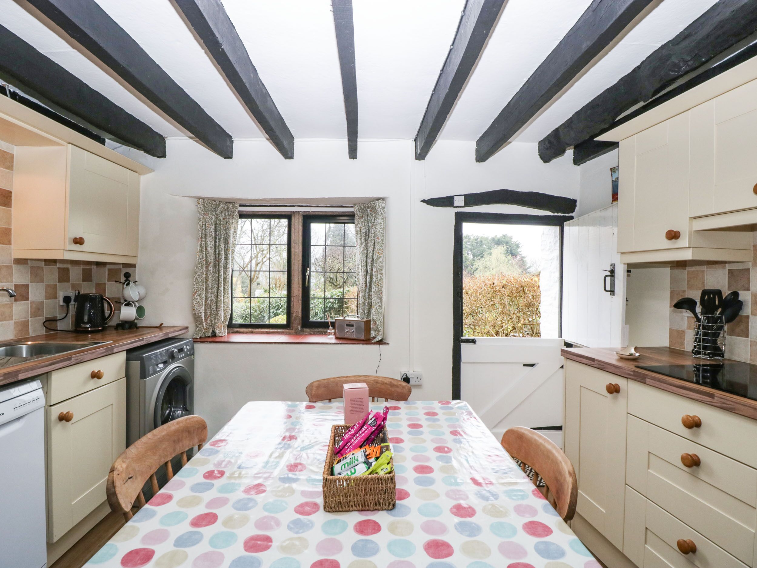 A compact kitchen with a dining area, featuring a window with curtains, a washing machine, and a table with a polka dot tablecloth.