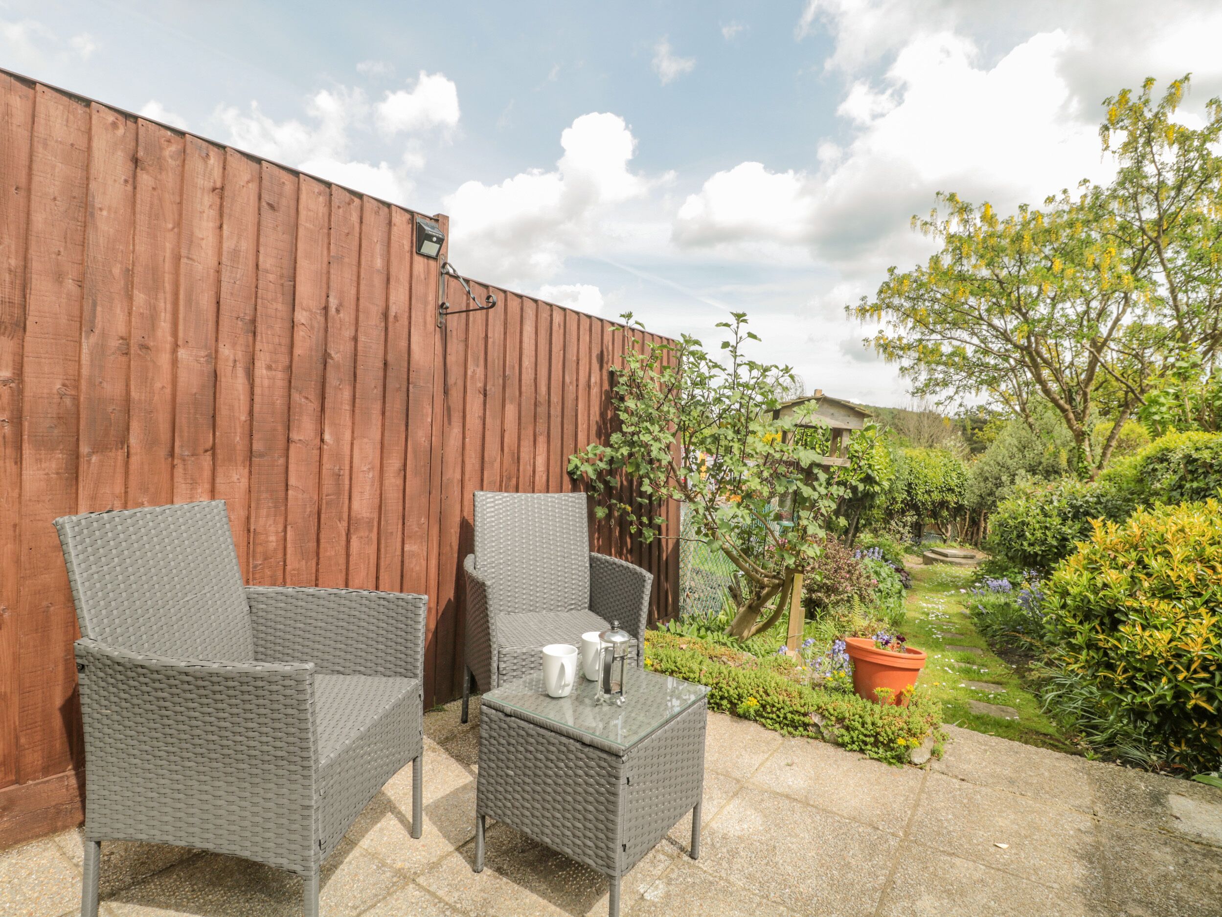 Een terras met rieten meubels en een tafel, omringd door een houten hek en een tuin met verschillende planten en een boom.