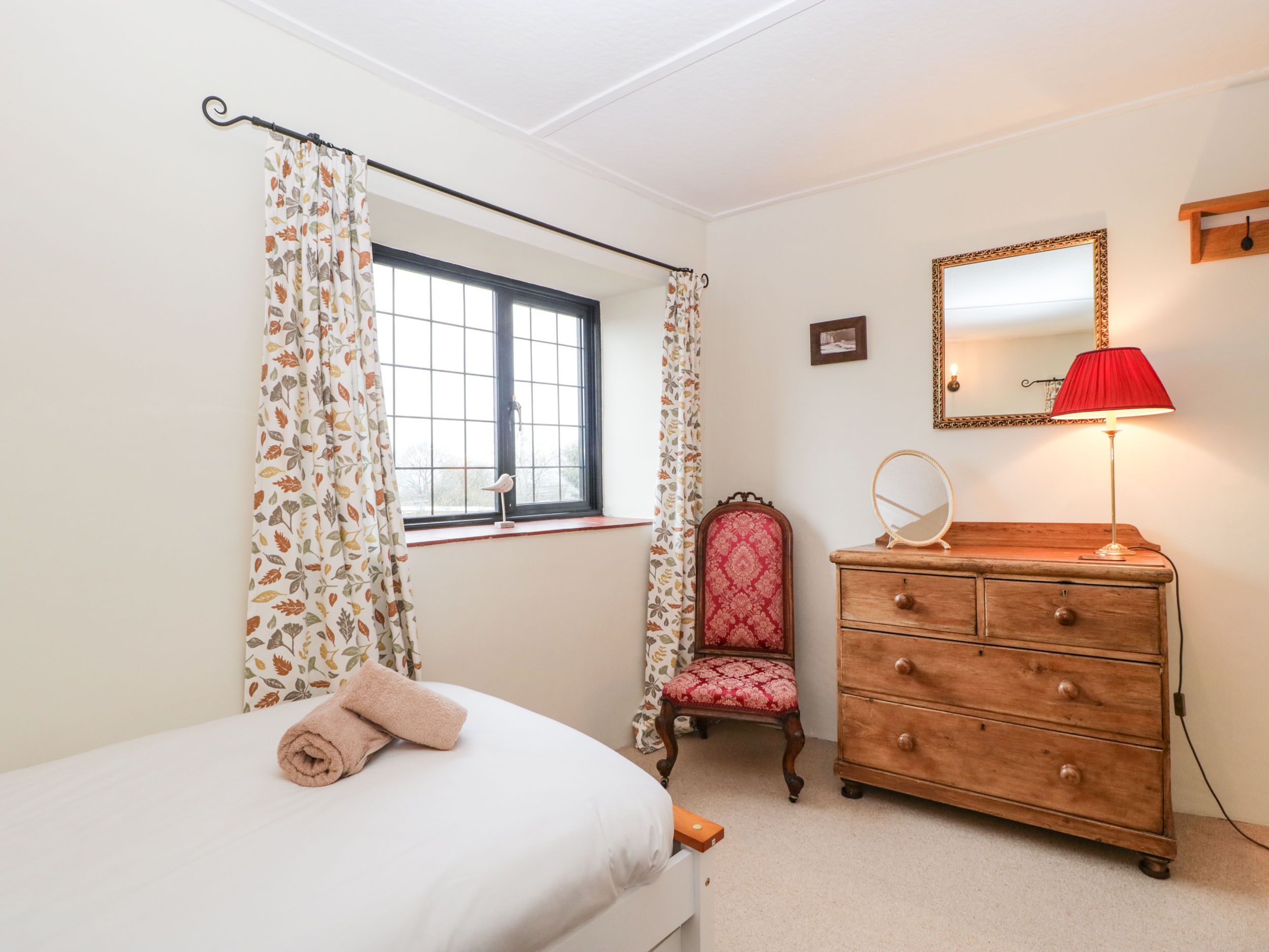 A bedroom with a bed, a dresser, a chair, a window with curtains, a mirror, and a lamp.