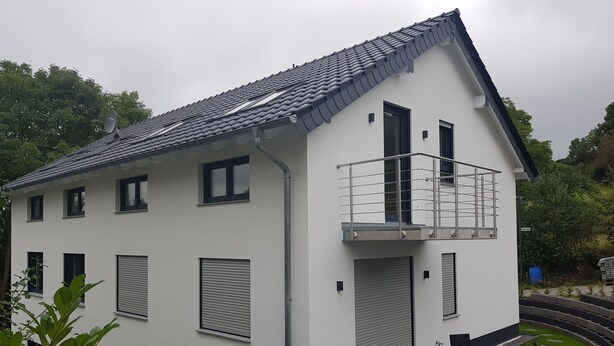 Un bâtiment moderne à deux étages, doté d’un toit plat, d’un balcon avec des rampes en métal et d’un petit patio comprenant une table et des chaises.