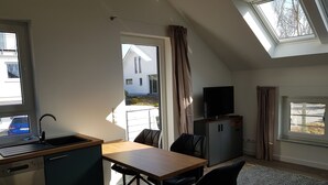 Une cuisine compacte équipée d’un lave-vaisselle, un coin repas avec une table et des chaises, un meuble gris avec un téléviseur, et une vue sur une maison par la fenêtre.