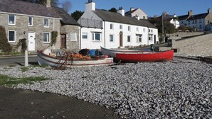 Un bâtiment situé sur la côte, doté d’une façade en pierre et d’une partie supérieure blanche, jouxtant une plage de galets où sont amarrés deux bateaux, l’un rouge et l’autre blanc.