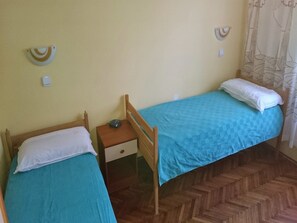 A room with two single beds, each with a wooden frame and a blue bedspread. There is a small wooden nightstand with a lamp and a power outlet between the beds. The walls are painted a light yellow, and there is a decorative wall hanging above the nightstand. - Apartment Nemo (Omisalj)