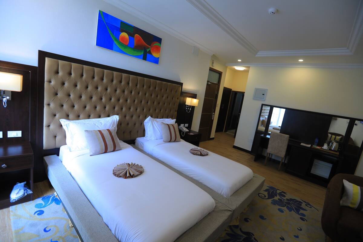 A hotel room with two beds, a large tufted headboard, a flat-screen TV, and a painting on the wall.