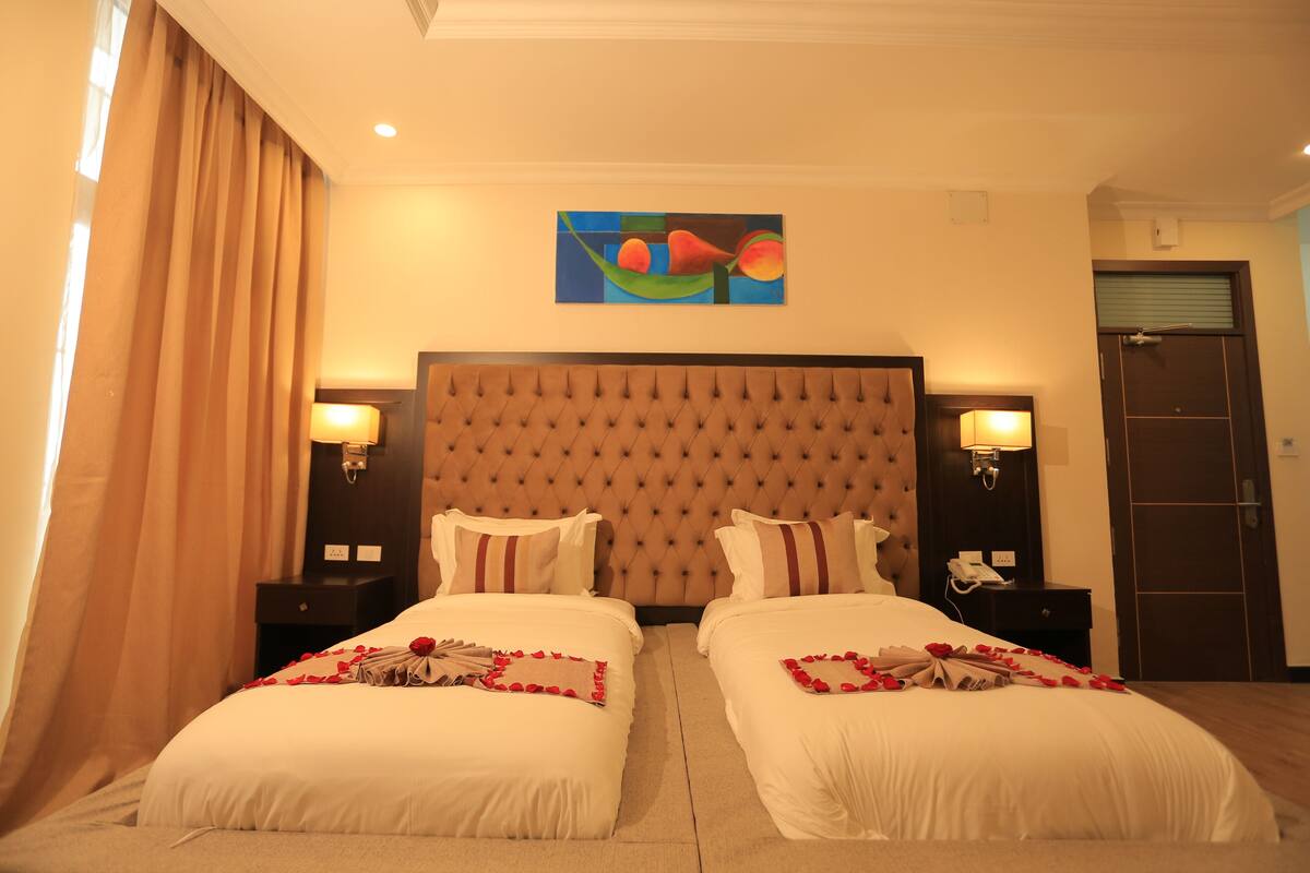 A hotel room with two beds, a large tufted headboard, bedside lamps, and a painting on the wall.