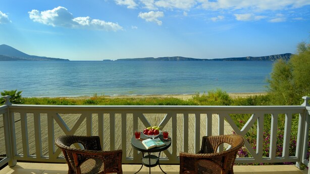 Un balcon avec des fauteuils en osier, une petite table avec des fruits et des boissons, et une vue sur une mer calme et des montagnes lointaines.