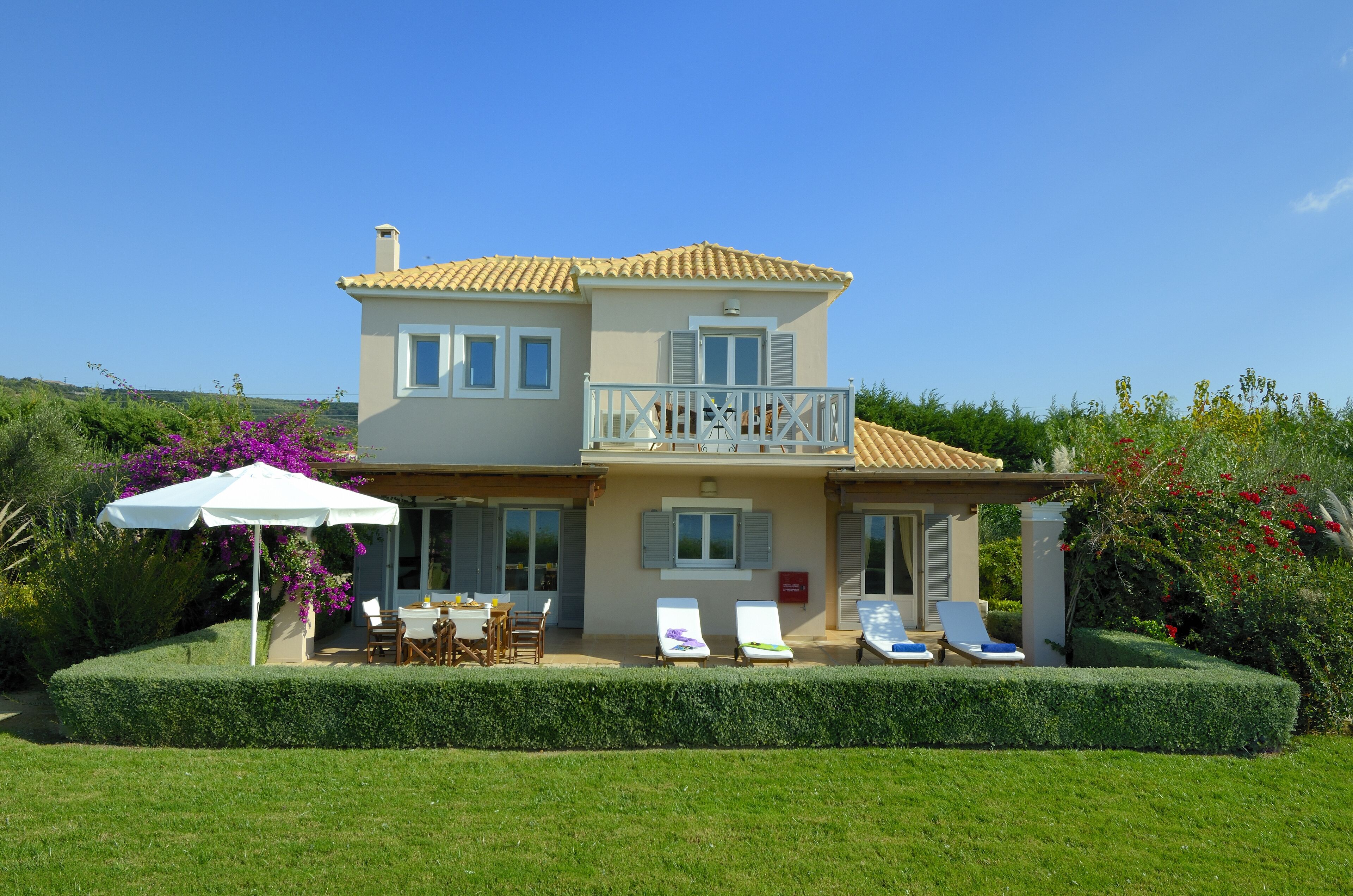 A two-story house with a balcony, a patio with a table and chairs, and a well-maintained garden.