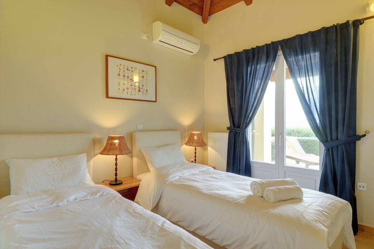 A hotel room with two beds, a window with blue curtains, an air conditioner, and a framed picture on the wall.