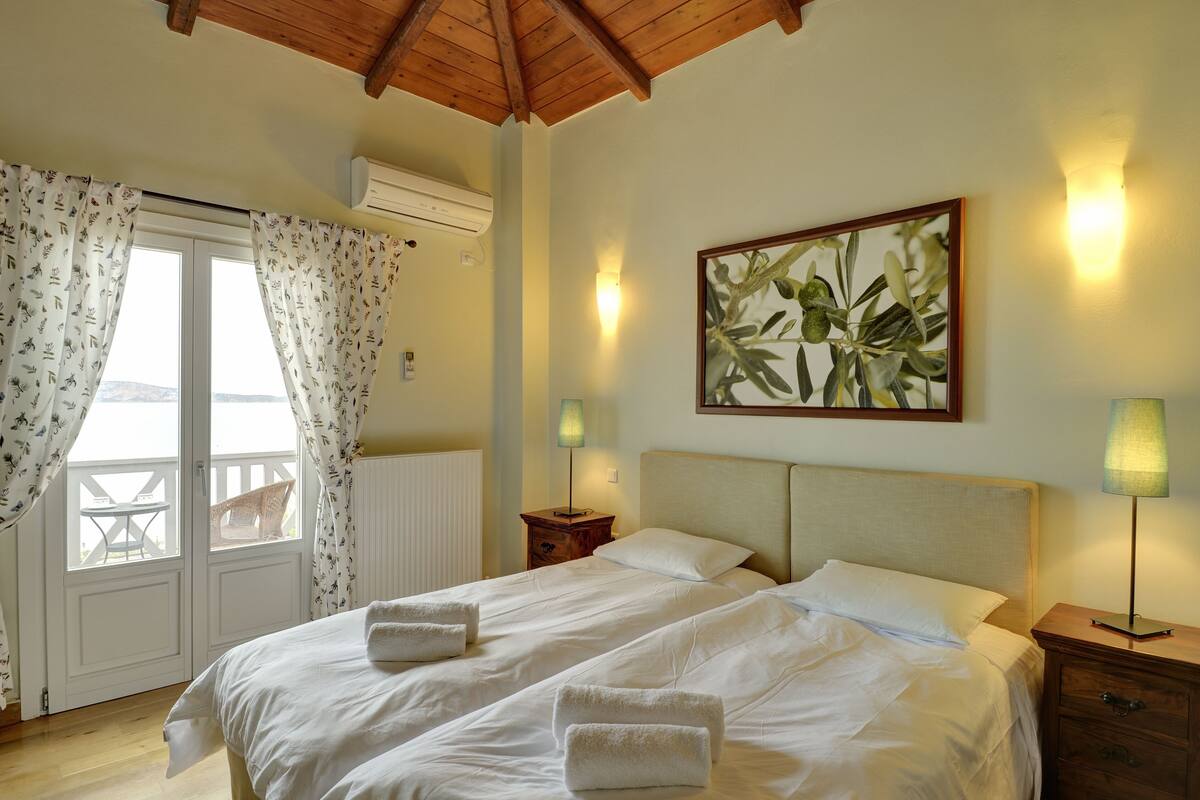 A hotel room with a bed, two bedside tables, a nightstand, a window with curtains, an air conditioning unit, and a painting on the wall.
