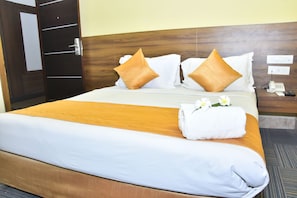 A neatly made bed with white and orange bedding, two decorative pillows, a rolled white towel, and a floral arrangement.