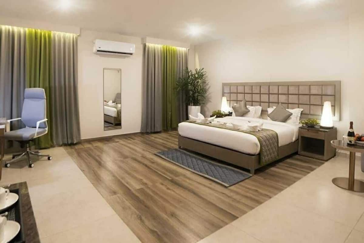 A modern hotel room with a large bed, a desk, a chair, and a small table with a lamp.