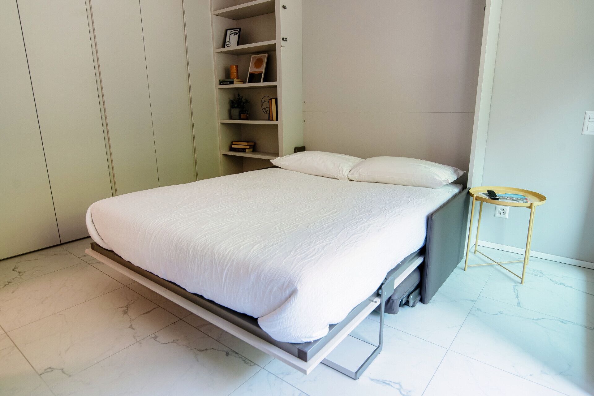 Een bed met een wit matras en kussens, een nachtkastje met een boek en telefoon, en een kledingkast met planken.