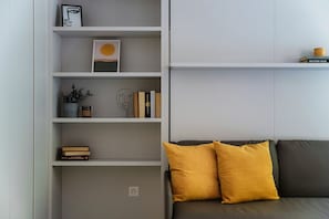 Un salon moderne avec un canapé gris, deux coussins de couleur jaune moutarde et une bibliothèque blanche contenant divers objets, notamment des livres et des objets décoratifs.