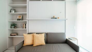 A modern living room with a grey sofa, two beige cushions, a white built-in shelving unit with books and decorative items, and a small round table with a remote control.