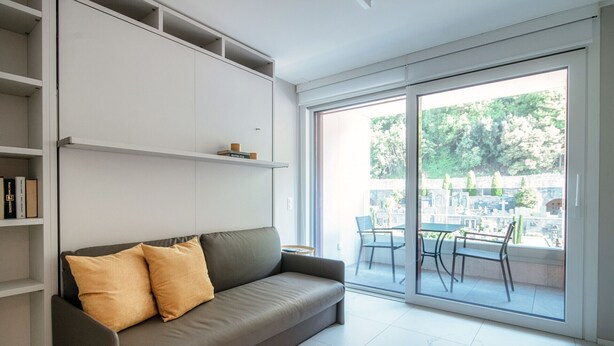 A modern living room with a grey sofa, two cushions, a bookshelf, and a large sliding glass door leading to an outdoor area with a table and chairs. - Studio with balcony near the center of Locarno (YELLOW STUDIO 12) (Locarno)