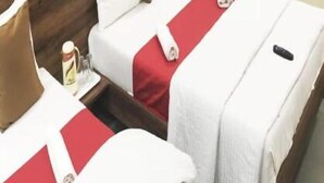 Habitación de hotel con dos camas, cada una con ropa de cama blanca y detalles en rojo, cabeceros de madera y mesitas de noche pequeñas con una botella y un control remoto.